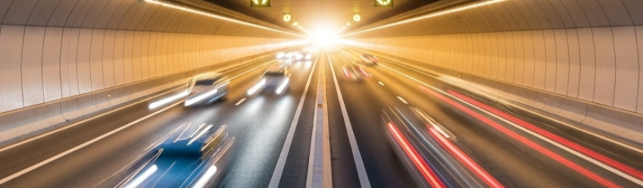 Das Bild zeigt eine moderne, mehrspurige Autobahn, die in einen Tunnel führt. Die Perspektive ist niedrig, als würde man aus einem fahrenden Auto schauen. Die Autobahn ist aus dunklem Asphalt und wird von dicken weißen und gelben Linien in mehrere Fahrspu